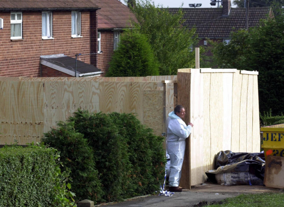 Person in forensic suit at a crime scene with wooden fence and house in background