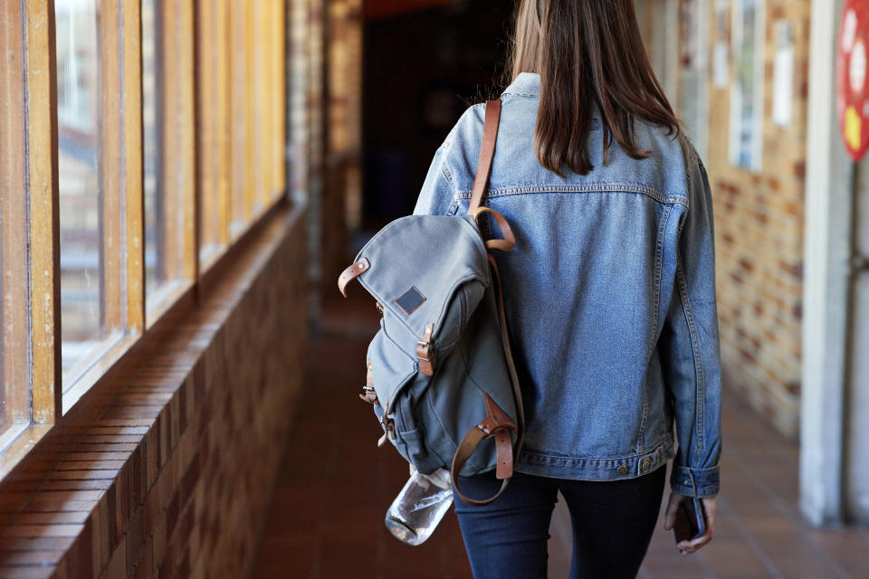 A person with a denim jacket and backpack is walking down a hallway, holding a phone. Their face is not visible.