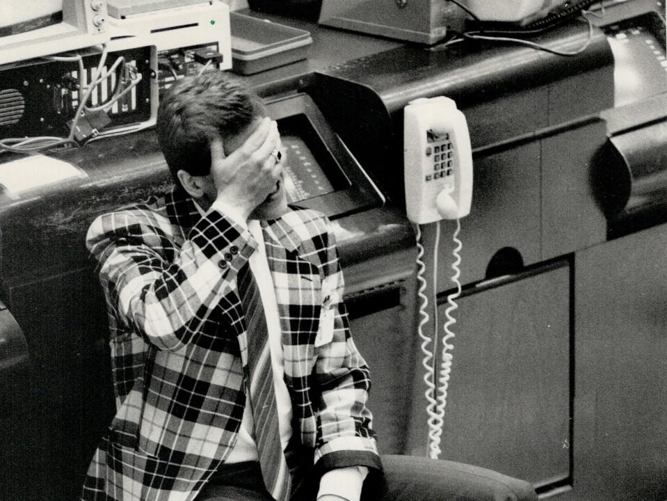 A black and white photo shows a man in a chechered vest covering his eyes while smoking a cigarette in the Toronto stock market. The floor is littered with paper.