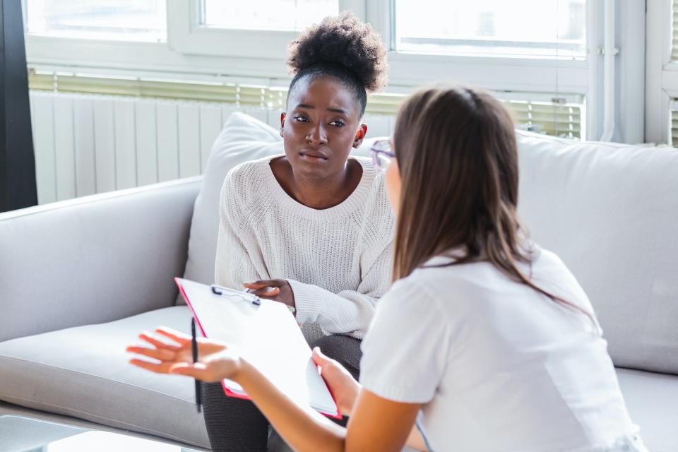 A woman sits on a couch as another woman speaks to her.