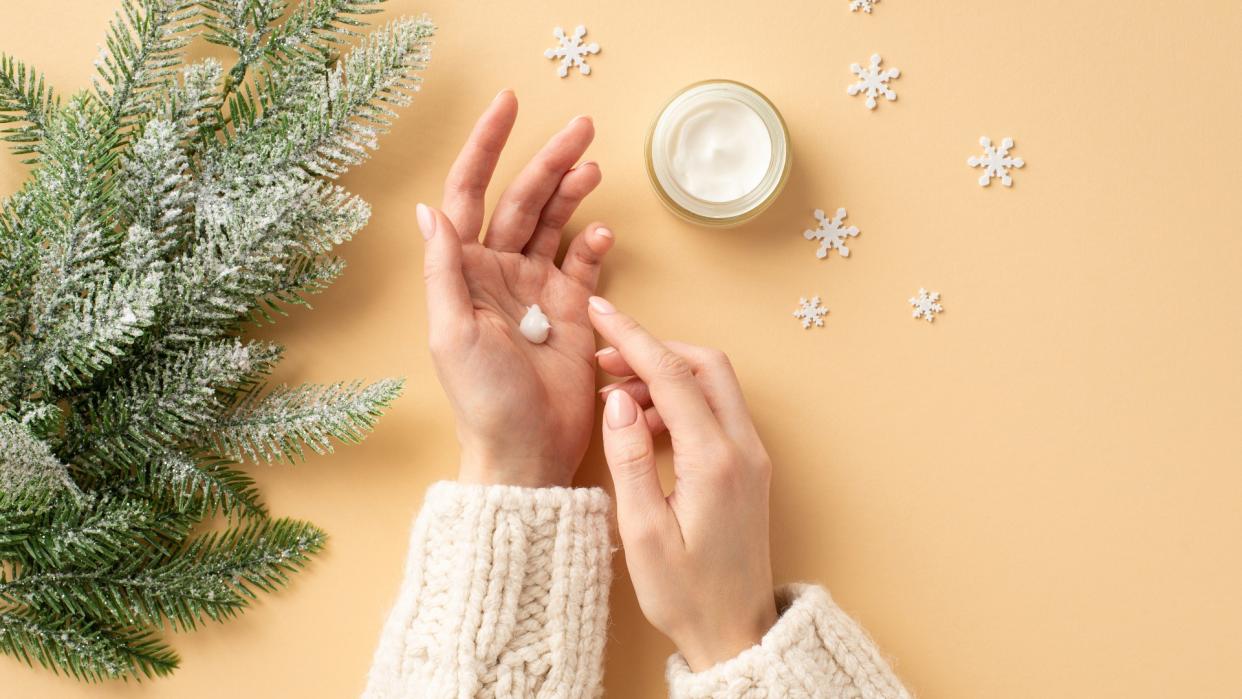  Girl's hands in white sweater applying cream on her hands from jar fir branches in frost and snowflakes on isolated pastel beige background . 