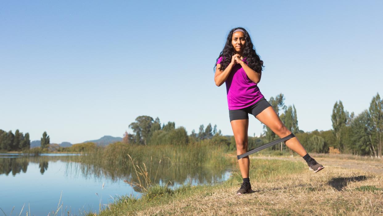  A woman stands at the edge of a lake, a resistance band around her lower legs, standing on one leg. 