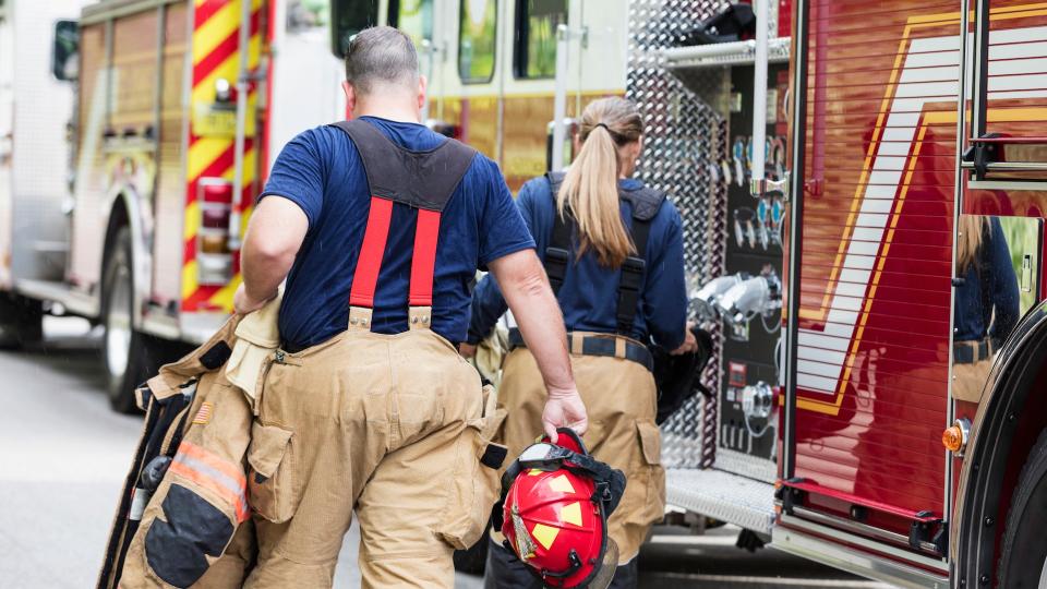 Rear view of two firefighters getting ready to answer a call, walking toward fire engines carrying their fire protection suits and helmets.