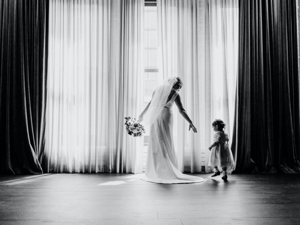 A bride holds out her hand for a flower girl as they walk to a window.