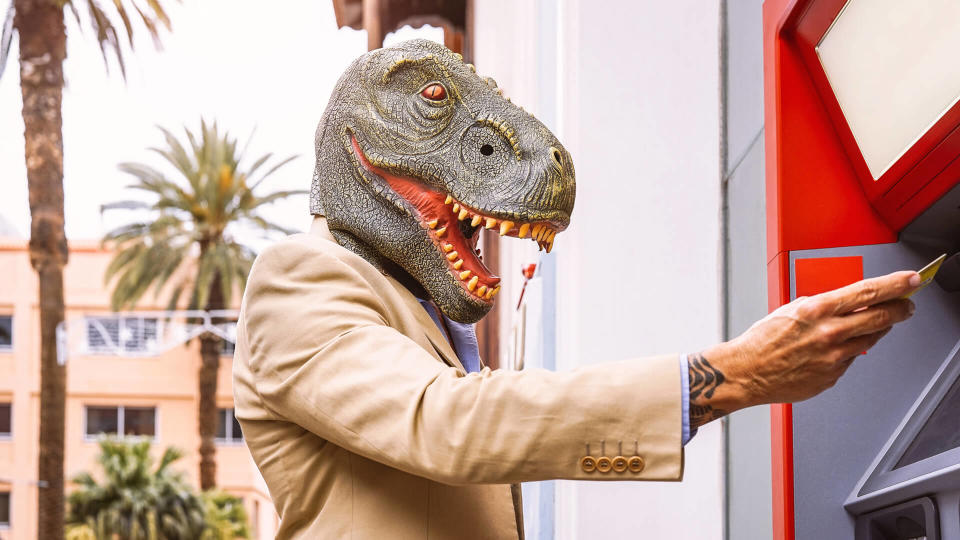 Senior man wearing t-rex dinosaur mask withdraw money from bank cash machine with debit card - Surreal image of half human and animal - Absurd and crazy concept of ATM advertise.