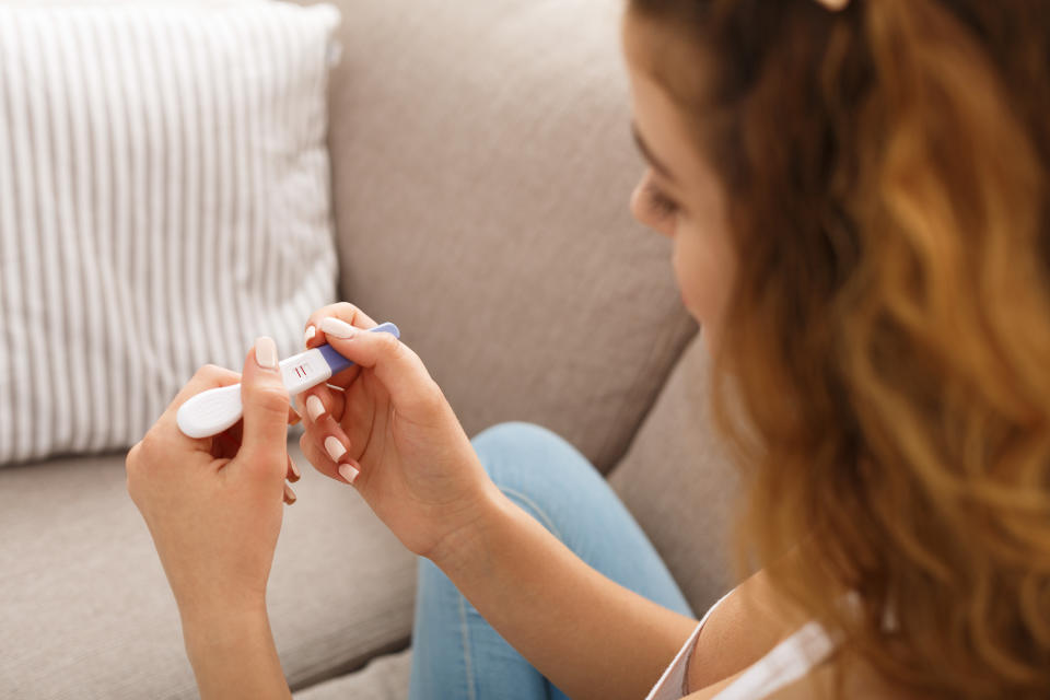 Worried redhead girl checking her recent pregnancy test, sitting on beige couch at home