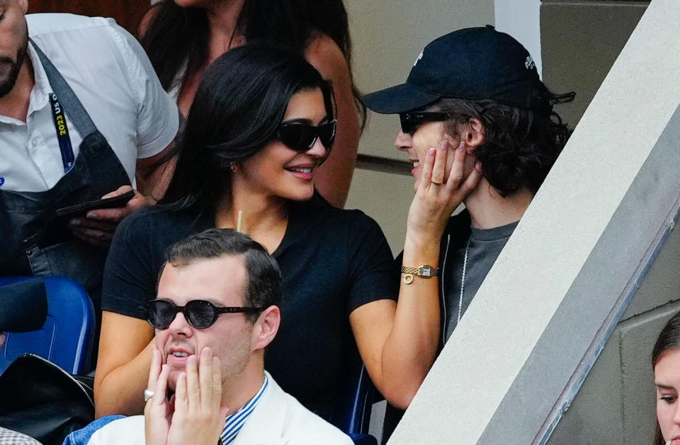 Kylie Jenner and Timothée Chalamet are smiling and interacting while seated at an event. Kylie is wearing sunglasses and a black top, and Timothée is wearing a cap