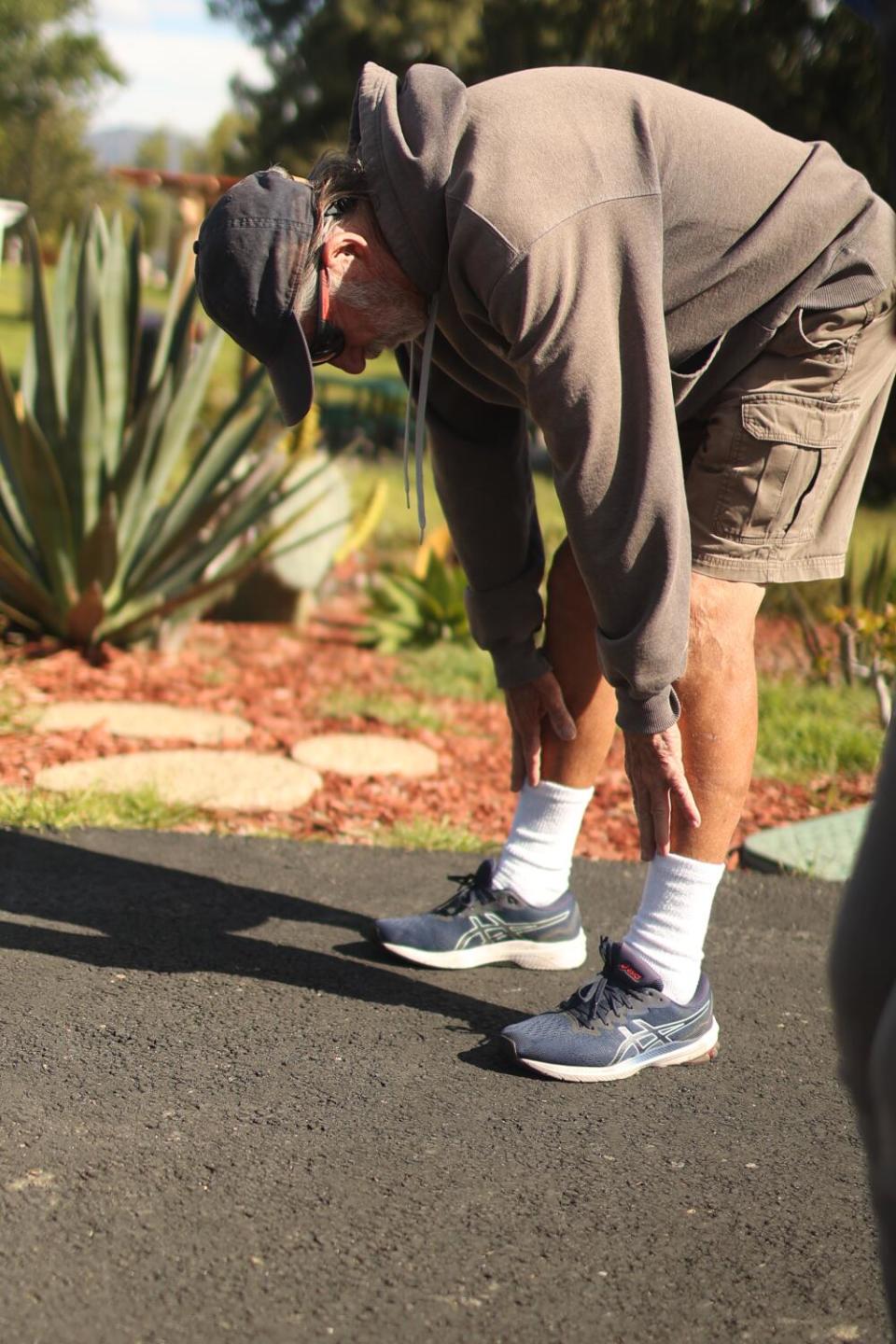 A man in a blue cap, brown hoodie and shorts bends at the waist and reaches for his toes