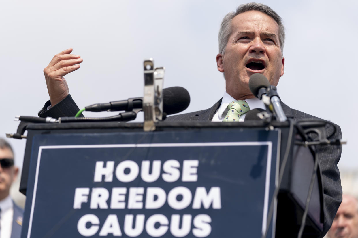 Rep. Jody Hice speaks into microphones at an outdoor podium with a plaque that reads House Freedom Caucus.