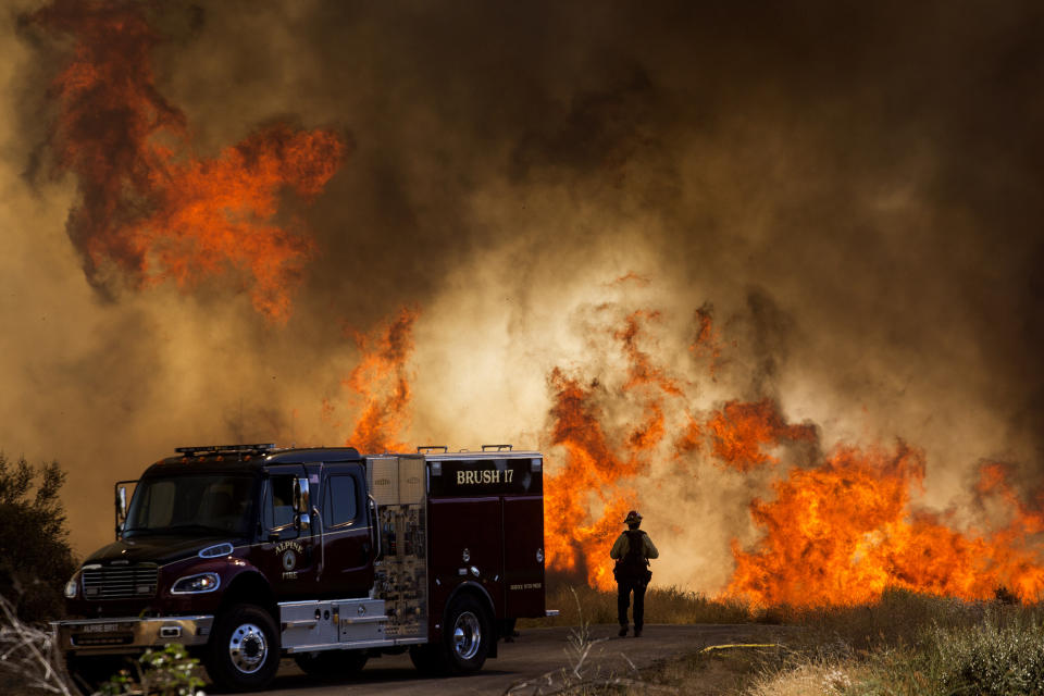 A firefighter stands next to a fire truck as flames flare at the Apple Fire in Cherry Valley, Calif., Saturday, Aug. 1, 2020. A wildfire northwest of Palm Springs flared up Saturday afternoon, prompting authorities to issue new evacuation orders as firefighters fought the blaze in triple-degree heat.(AP Photo/Ringo H.W. Chiu)