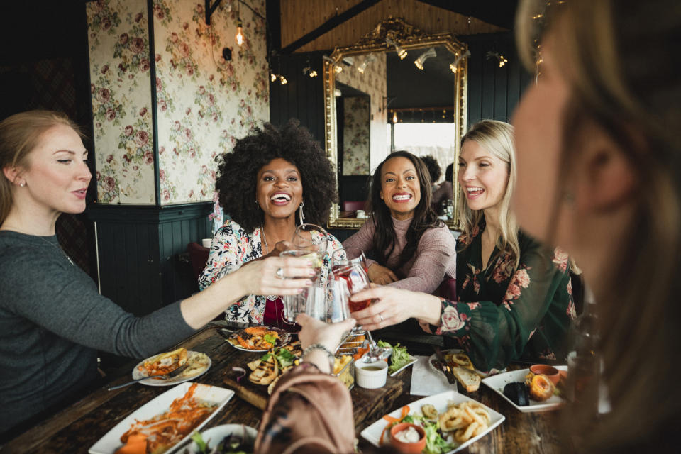 A group of five women, including Kim Kardashian, are seated at a restaurant table, laughing and raising their drinks in a toast