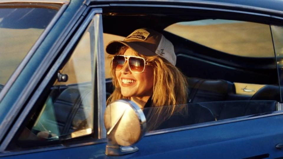 natasha lyonne drives away in blue car wearing a trucker hat