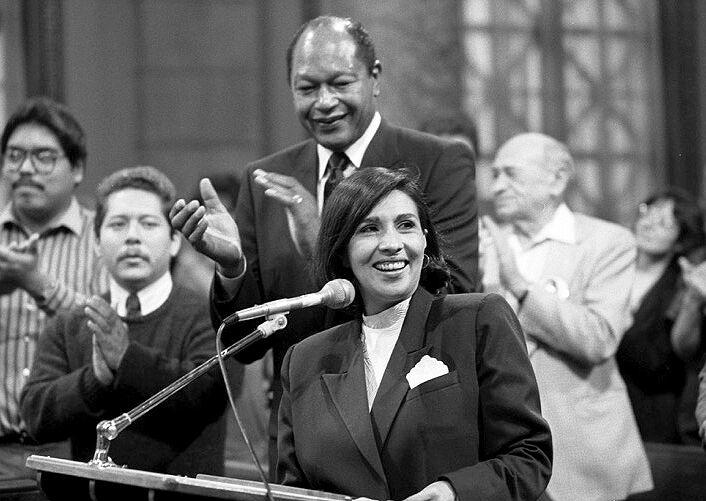 A black-and-white photo of Gloria Molina speaking into a microphone, smiling, and Tom Bradley behind her applauding.