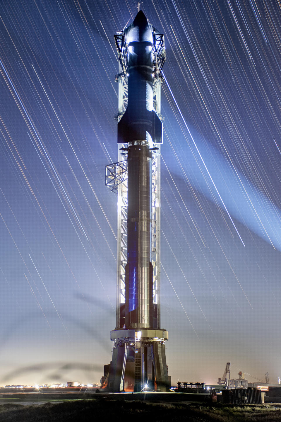 A large stainless steel rocket stands on the launchpad. the finned top half is covered in heat tiles. The streaks of stars from long camera exposures line the night sky.