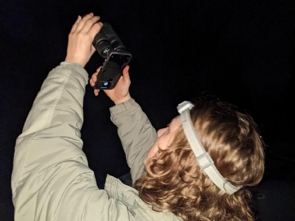 woman wearing headlamp and thick jacket holding phone up to binoculars