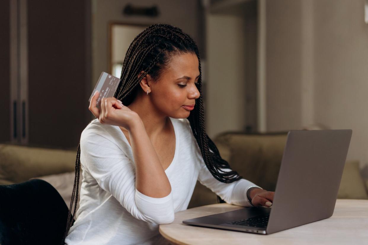 Black woman with knotless braids holding credit card sitting at table with laptop online shopping in living room