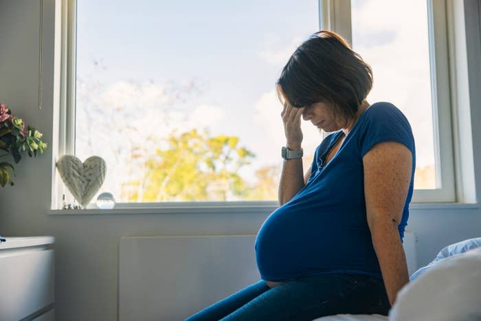 A pregnant woman sitting on a bed, holding her head in apparent discomfort, near a window with daylight streaming in