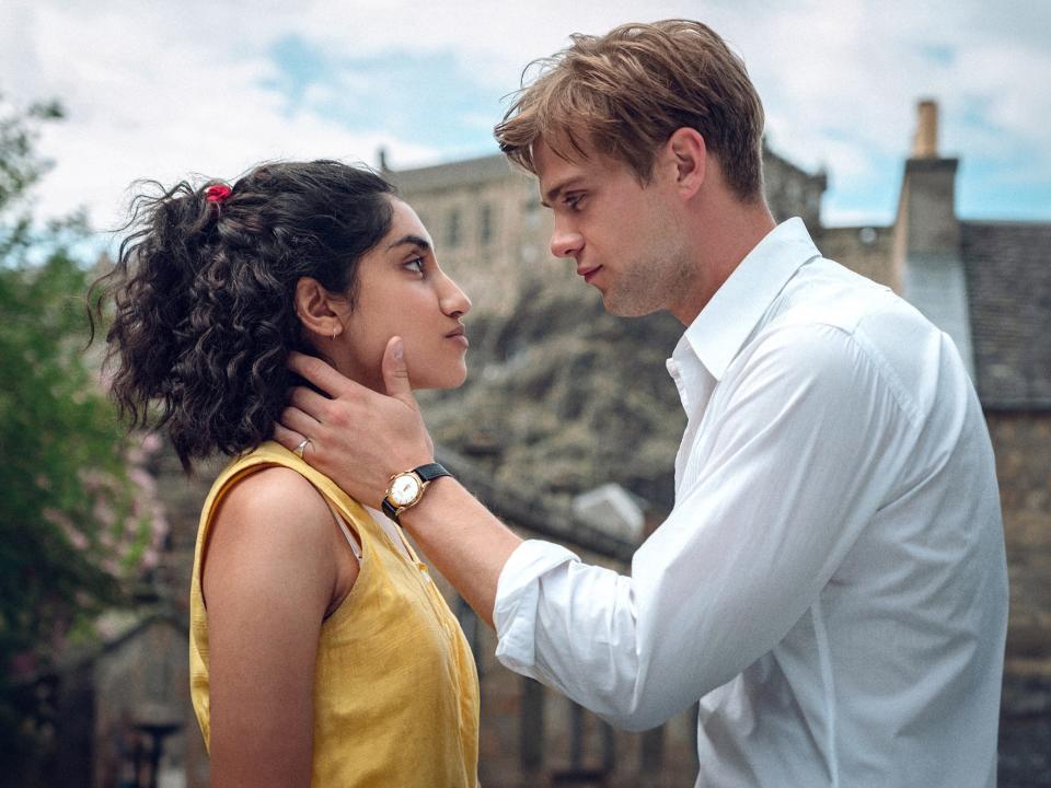 a young man cradles a woman's neck and jaw as they look intensely into each others' eyes face to face, standing outdoors in a picturesque village