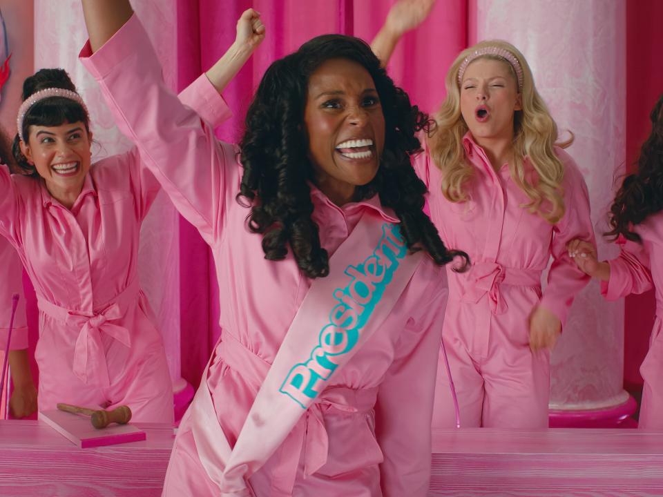 Issa Rae in a scene from "Barbie" where President Barbie is raising her hand and cheering with a room of other Barbies.