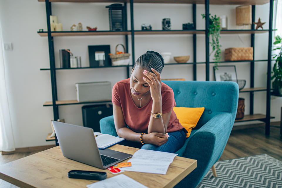 A woman sits on a blue couch, looking stressed while reviewing papers in front of a laptop in a home office setting