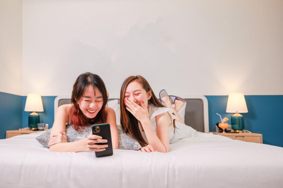 Two Asian females' enjoys using their smartphone and taking their photo on the bed.