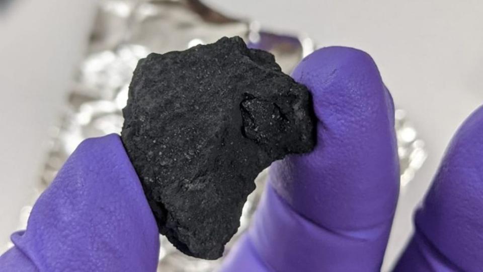 a chunk of carbonaceous chondrite meteorite is held between a gloved finger and thumb; the meteorite resembles black coal