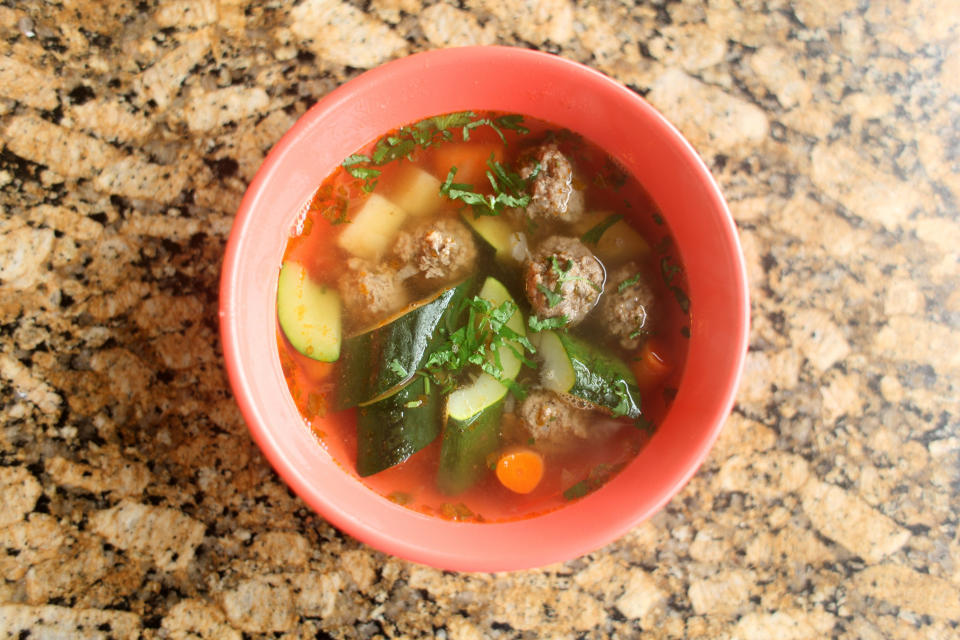 A bowl of hearty vegetable and meatball soup, containing zucchini, carrots, and cilantro, on a textured stone surface