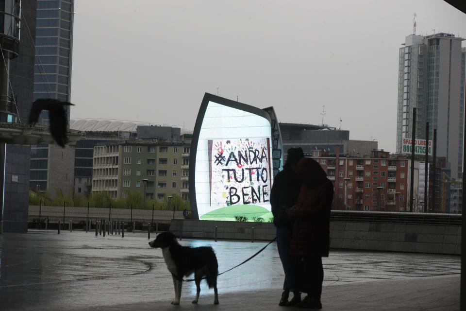 In this photo taken on Saturday, March 14, 2020 a couple and their dog stand by a billboard reading in Italian "Everything will be alright", in Milan, Italy. The nationwide lockdown to slow coronavirus is still early days for much of Italy, but Italians are already are showing signs of solidarity. This week, children’s drawings of rainbows are appearing all over social media as well as on balconies and windows in major cities ready, ‘’Andra’ tutto bene,’’ Italian for ‘’Everything will be alright.’’ For most people, the new coronavirus causes only mild or moderate symptoms. For some, it can cause more severe illness, especially in older adults and people with existing health problems. (AP Photo/Luca Bruno)