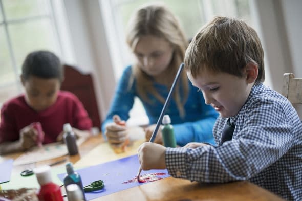 Children in a family home.  Three children sitting at a table using glue and paint to create decorations.