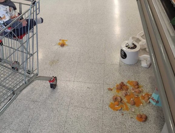 A toddler sits in a shopping cart near a spill on the floor involving broken eggs and a toilet paper roll