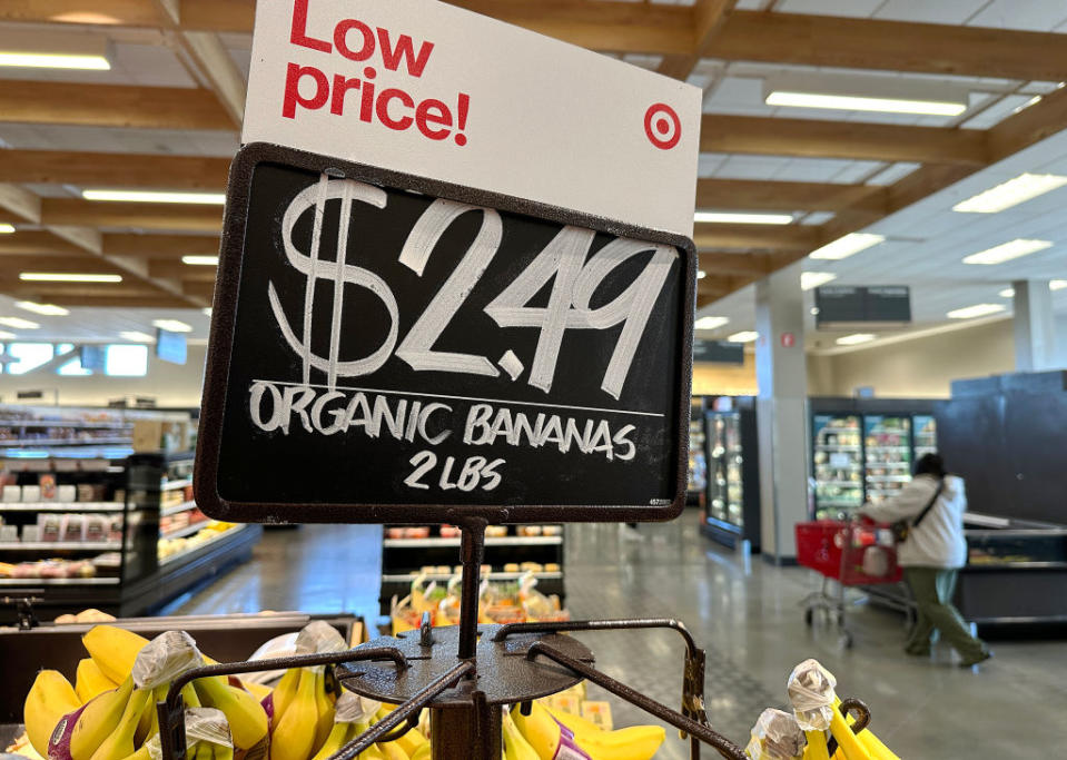 A Target price sign reads "$2.49 ORGANIC BANANAS 2 lbs" hanging above bananas in a store; there's a shopper with a cart in the background