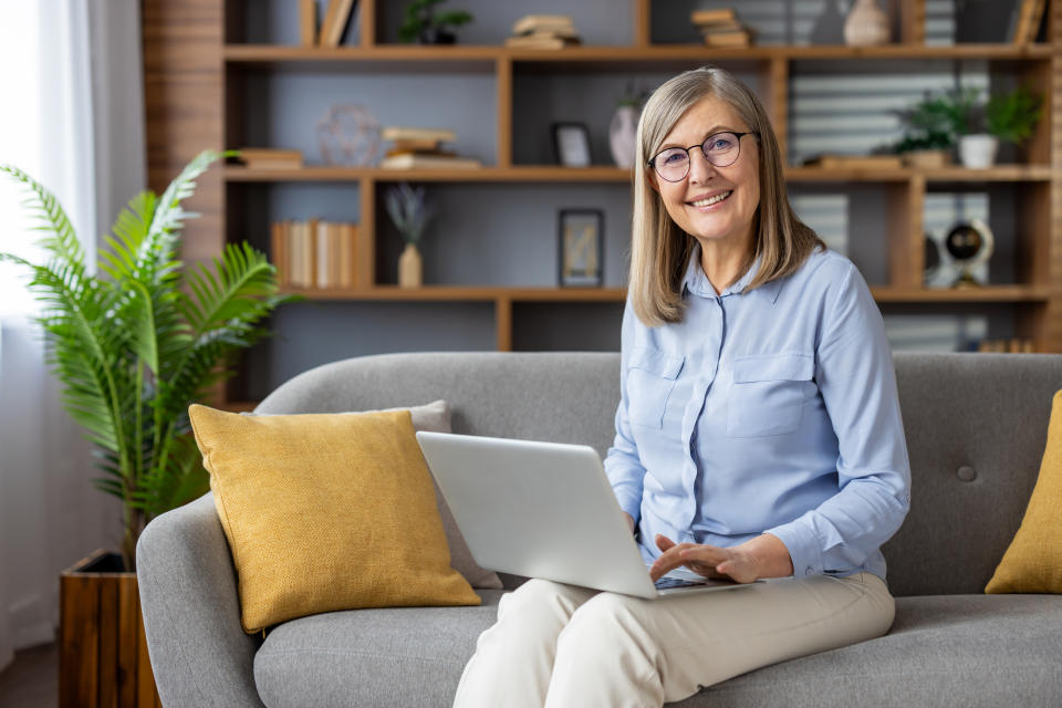 An older woman with glasses and a blue blouse is sitting on a gray couch with a laptop on her lap. Bookshelves and plants are in the background