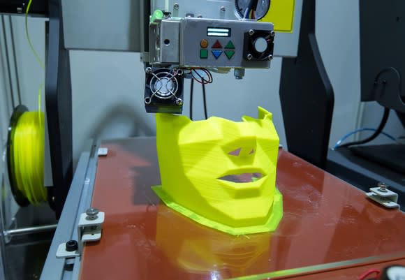 A 3D printer printing a human face or mask from yellow plastic.