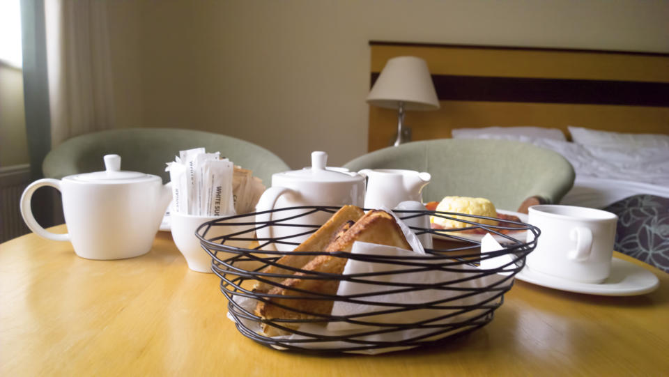 Breakfast tray with teapots, cups, sugar packets, and pastries on a table in a cozy hotel room. Two green chairs and a bed with pillows are in the background