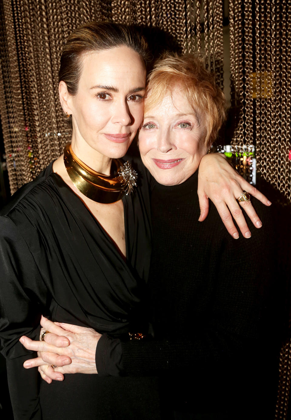 Two women embracing, one wearing a black turtleneck and the other wearing a draped blouse with a statement necklace