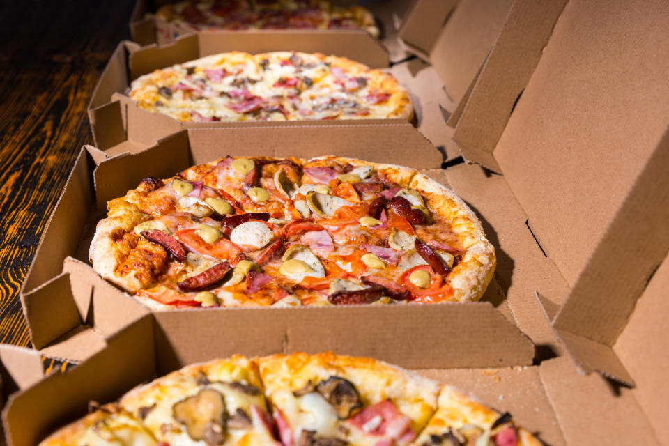 Several open pizza boxes containing various pizzas with different toppings arranged in a row on a wooden surface