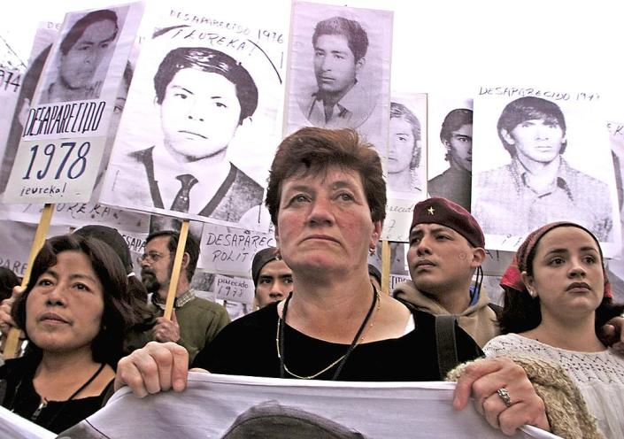 Familiares de personas desaparecidas durante el periodo de contrainsurgencia en México marchan el 2001. Se ve a siete personas portando carteles con las  fotos de hombres jóvenes que dicen 