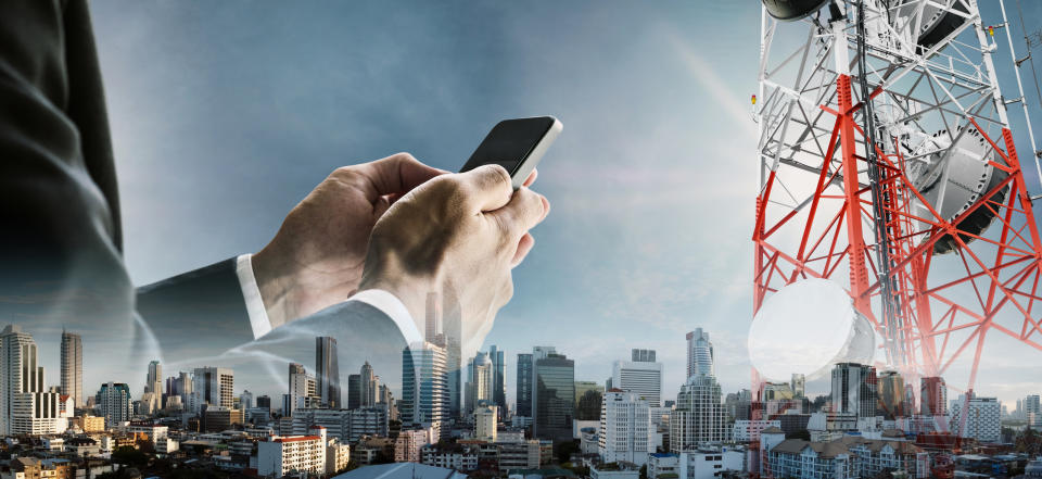 Superimposed over a city skyline, a red and white cell tower and a businessman using a smartphone.