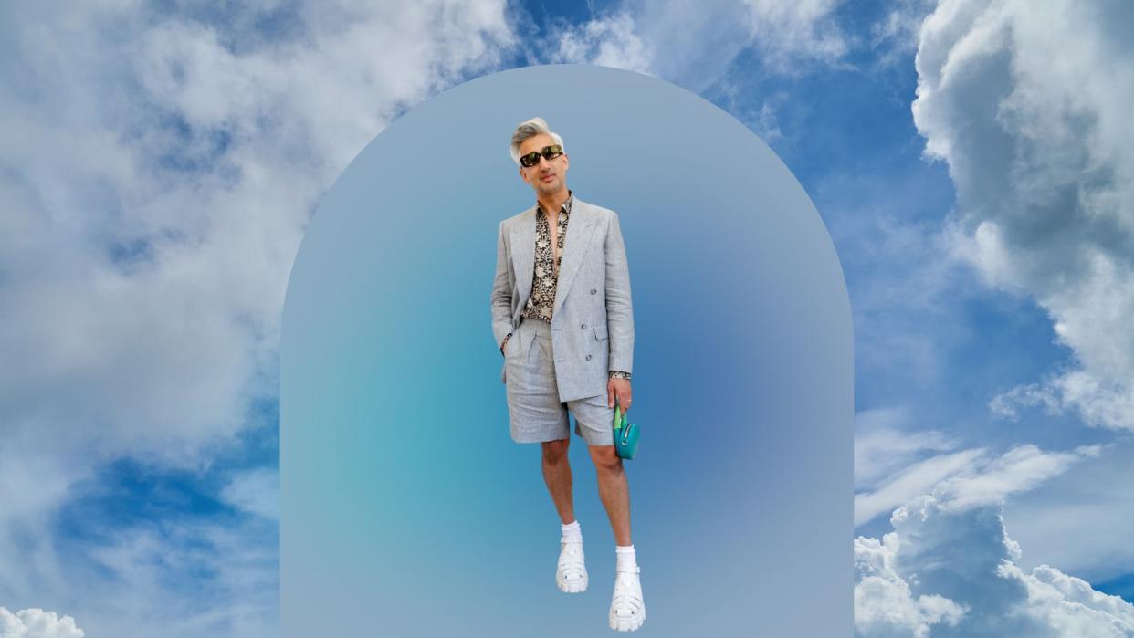  Tan France in a grey suit with shorts and sunglasses on a sky blue background. 