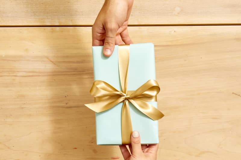 Overhead shot of someone handing over a small package wrapped in light blue paper, tied in a gold bow.