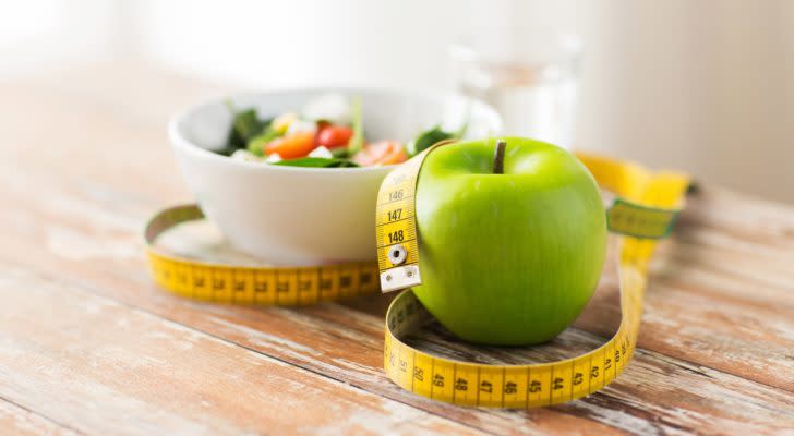 a bowl of vegetables, a green apple, and a measuring tape sit on a wooden table