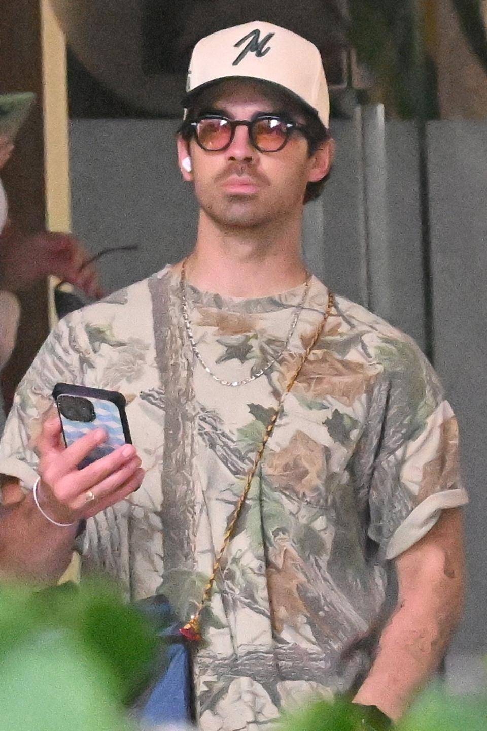 Joe Jonas in camo tee and cap holds phone, sporting sunglasses and a necklace