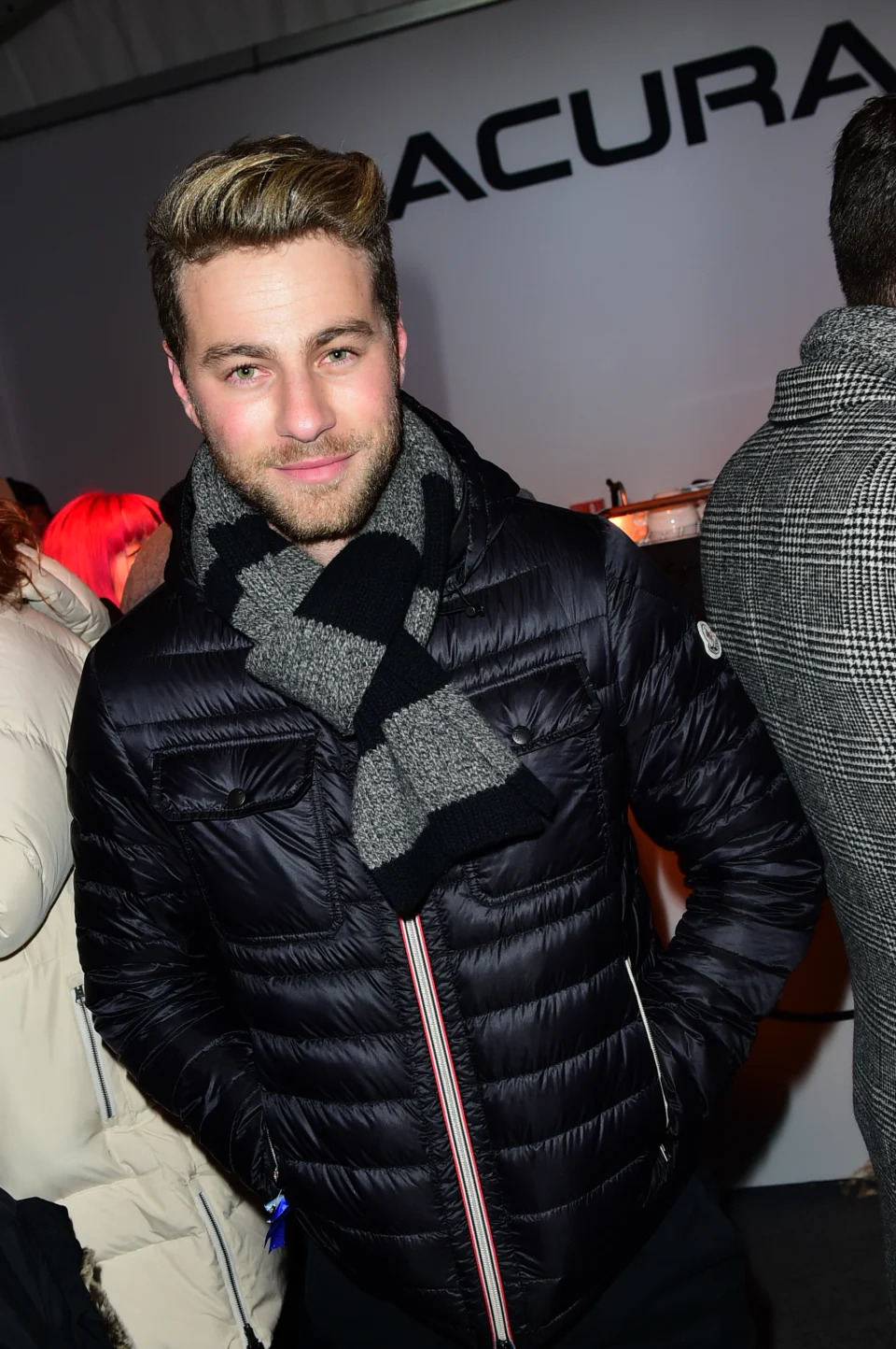 A man in a black puffer jacket and striped scarf smiles at an event. The background has part of the word "ACURA" visible