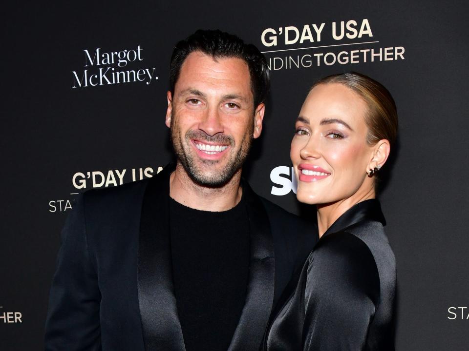 A man and woman (Maksim Chmerkovsky and Peta Murgatroyd) stand smiling for the camera in all-black outfits at a red carpet event.