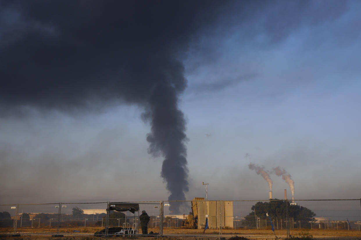 An Israeli soldier stands guard next to an Iron Dome air defense system as smoke rises from an oil tank on fire after it was hit by a rocket fire from Gaza Strip, near the town of Ashkelon, Israel,, Wednesday, May 12, 2021. (AP Photo/Ariel Schalit)