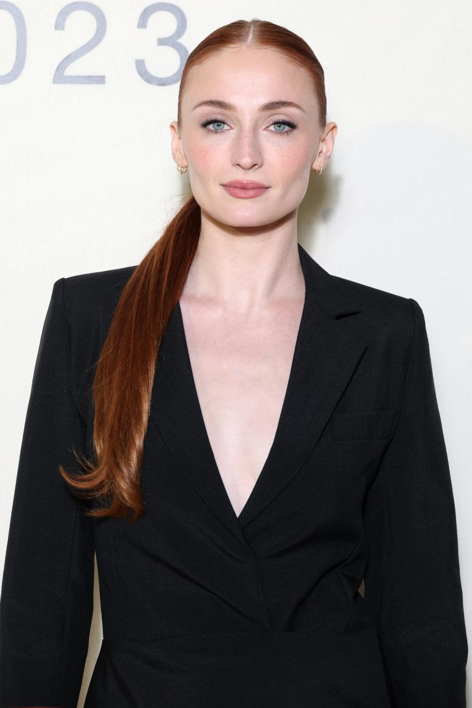 Sophie Turner poses in a black blazer, standing against a numbered backdrop