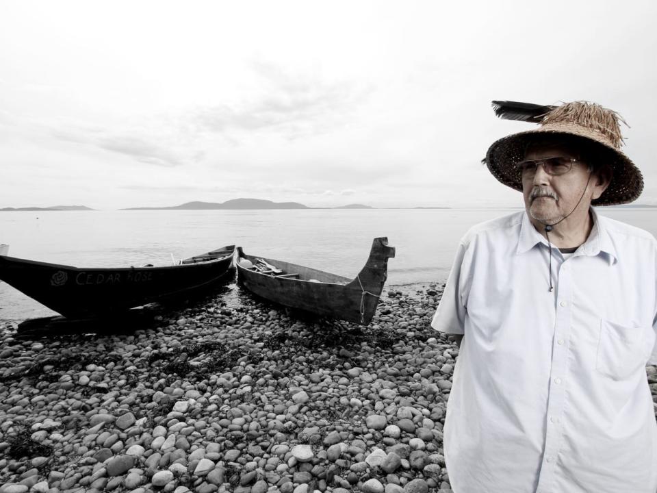 Chief Bill James, Lummi Nation. A man poses for a portrait in front of a canoe and water.