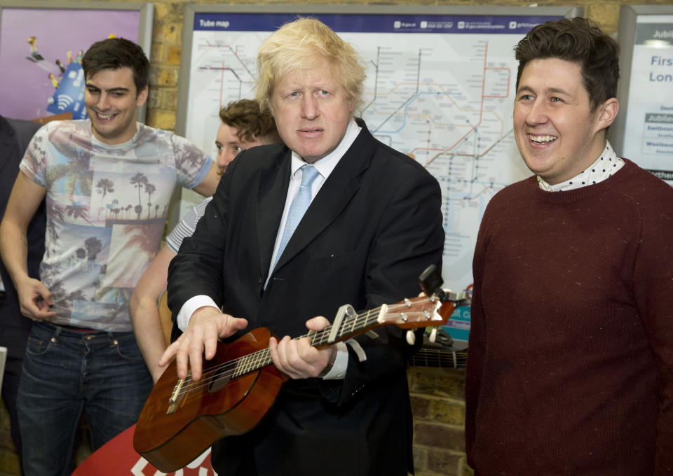 FILE - In this Monday, March 23, 2015 file photo Mayor of London Boris Johnson plays with a guitar in front of the busking band The Tailormade during a photocall to promote two new schemes aimed at supporting and promoting busking and street performance, at London Bridge Station in London. (AP Photo/Alastair Grant, File)