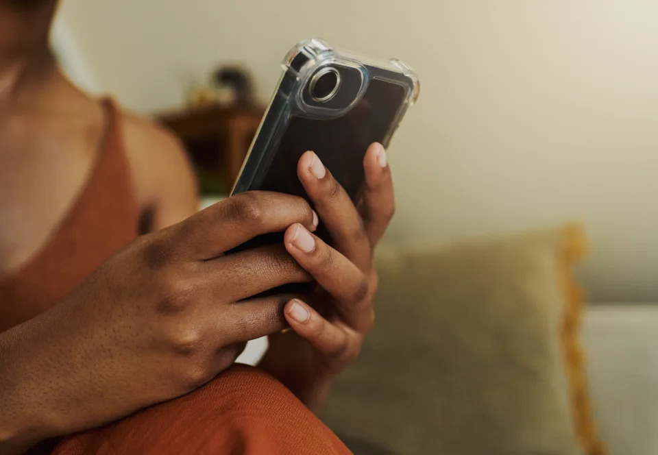 Wanita muda kulit hitam yang tidak dikenali, duduk dengan nyaman di sofa sambil mengirim pesan teks di telepon genggamnya sambil berpakaian santai dengan ruang fotokopi, stok foto.