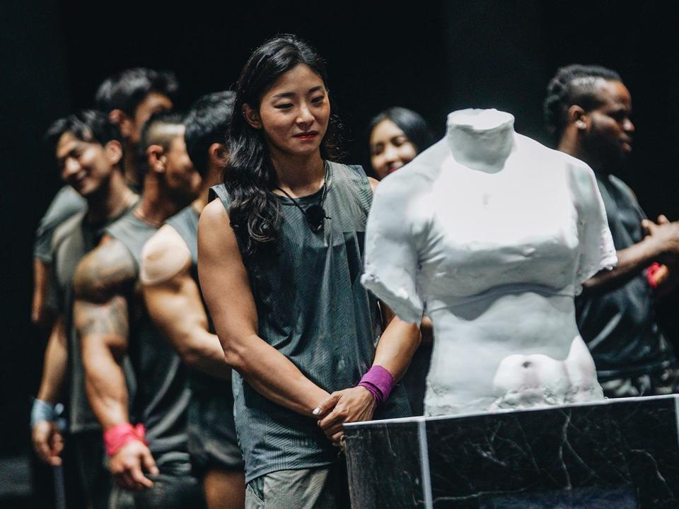 jang eun-sil on physical 100, standing in front of a plaster cast of her own torso with her hands folded in front of her. while the contestants standing behind her are chatting, jang appears reflective